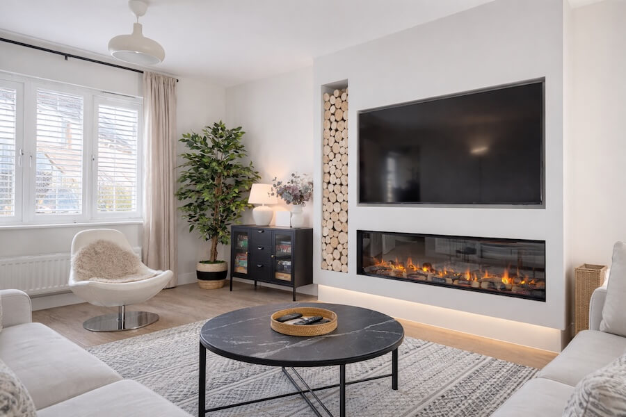 Living room with dark media wall, large wall-mounted TV, electric fire beneath, hexagonal wooden wall art, white armchair with sheepskin throw, and tall indoor plant