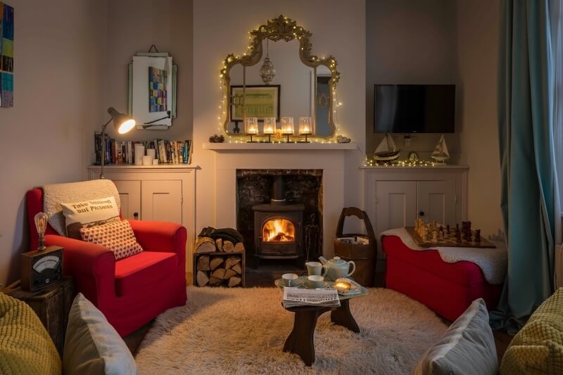 Cosy living room with roaring wood-burning stove, red armchairs, tea set, and chessboard in a Camber Sands holiday cottage – ideal for your winter escape.