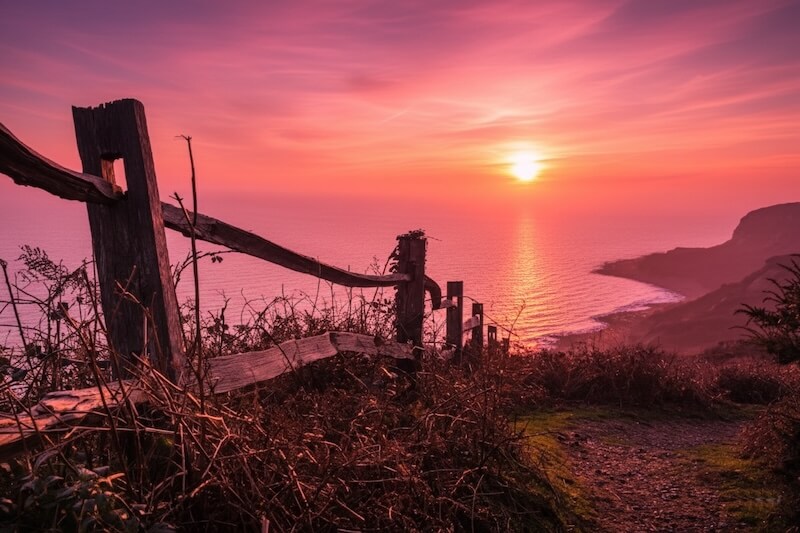 Dramatic pink and orange winter sunset viewed from a wooden fence along the clifftop path at Firehills in Hastings Country Park, overlooking the sea – perfect for invigorating walks during your winter escape in Camber Sands.